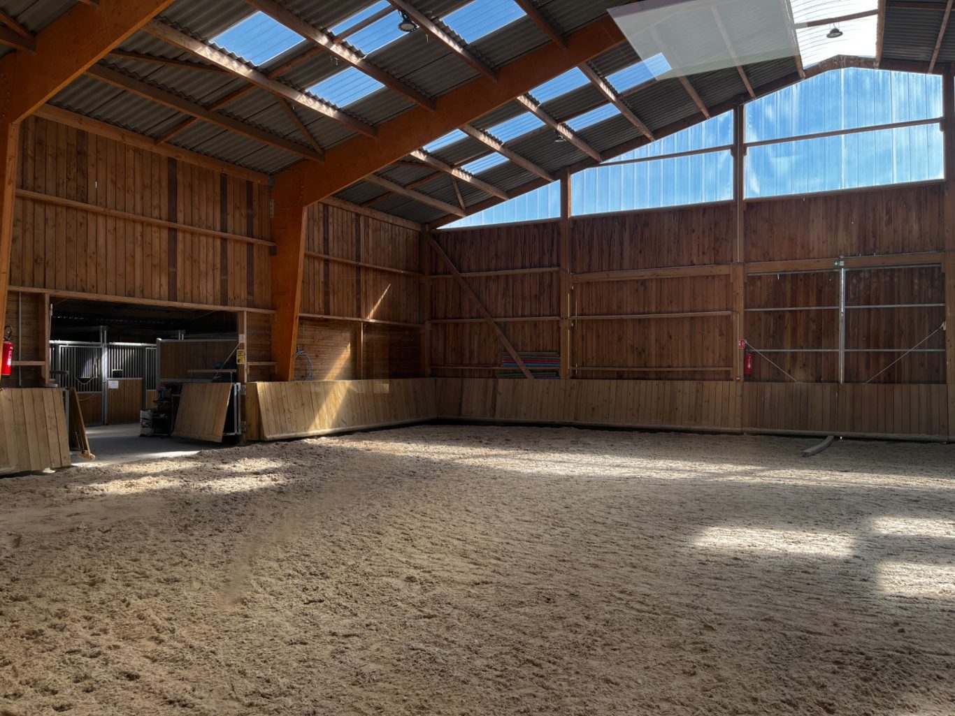 Manège de 20m x 18m Salle de manège spacieuse avec un sol en sable et des murs en bois. Lumière naturelle entrante.